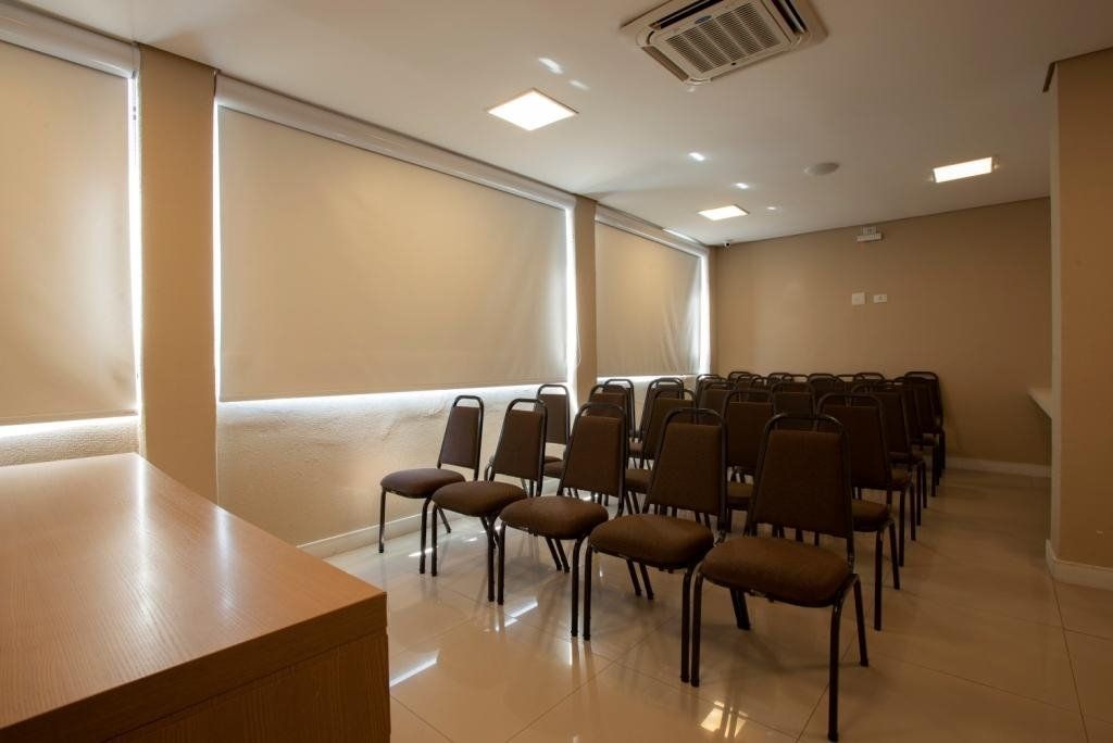 Sala de conferências vazia com fileiras de cadeiras, paredes bege e uma mesa.