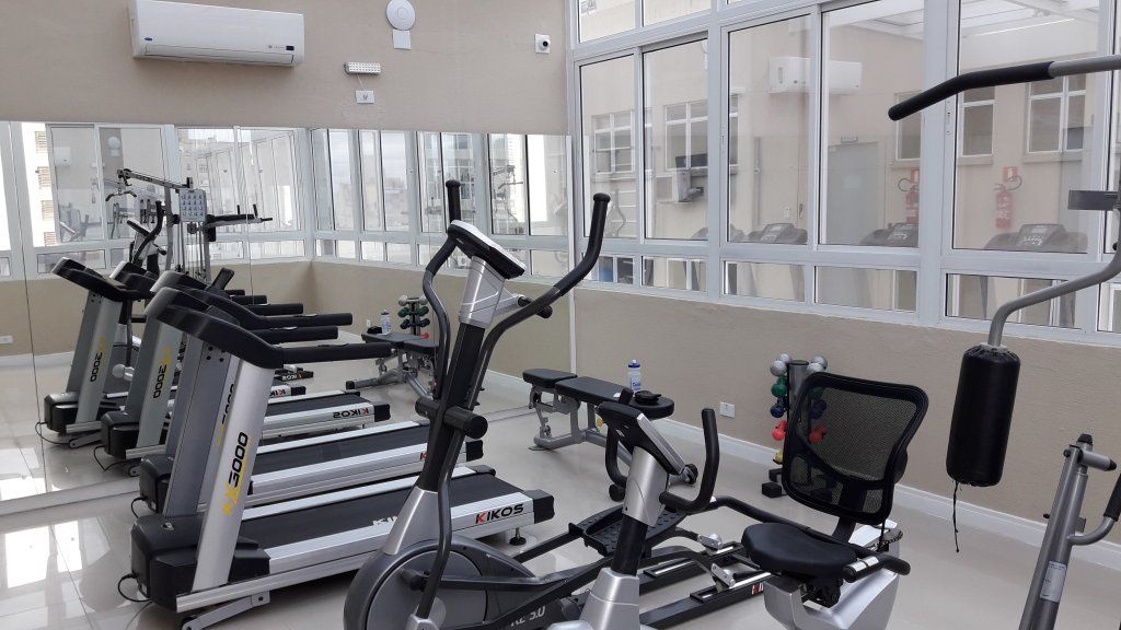 Academia com esteiras, elíptico, pesos e espelhos. As janelas oferecem luz natural.