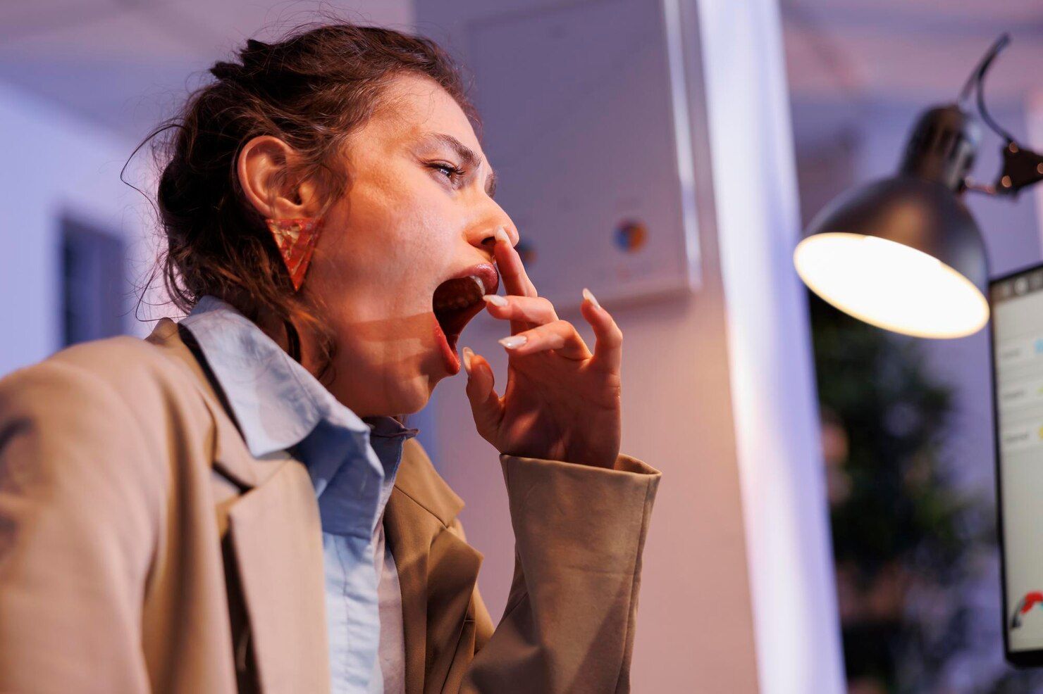 A woman is yawning while sitting at a desk in front of a computer.