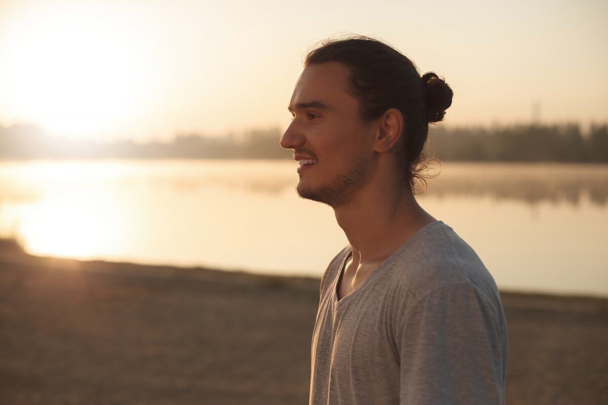 A man with a bun is standing on the beach at sunset.