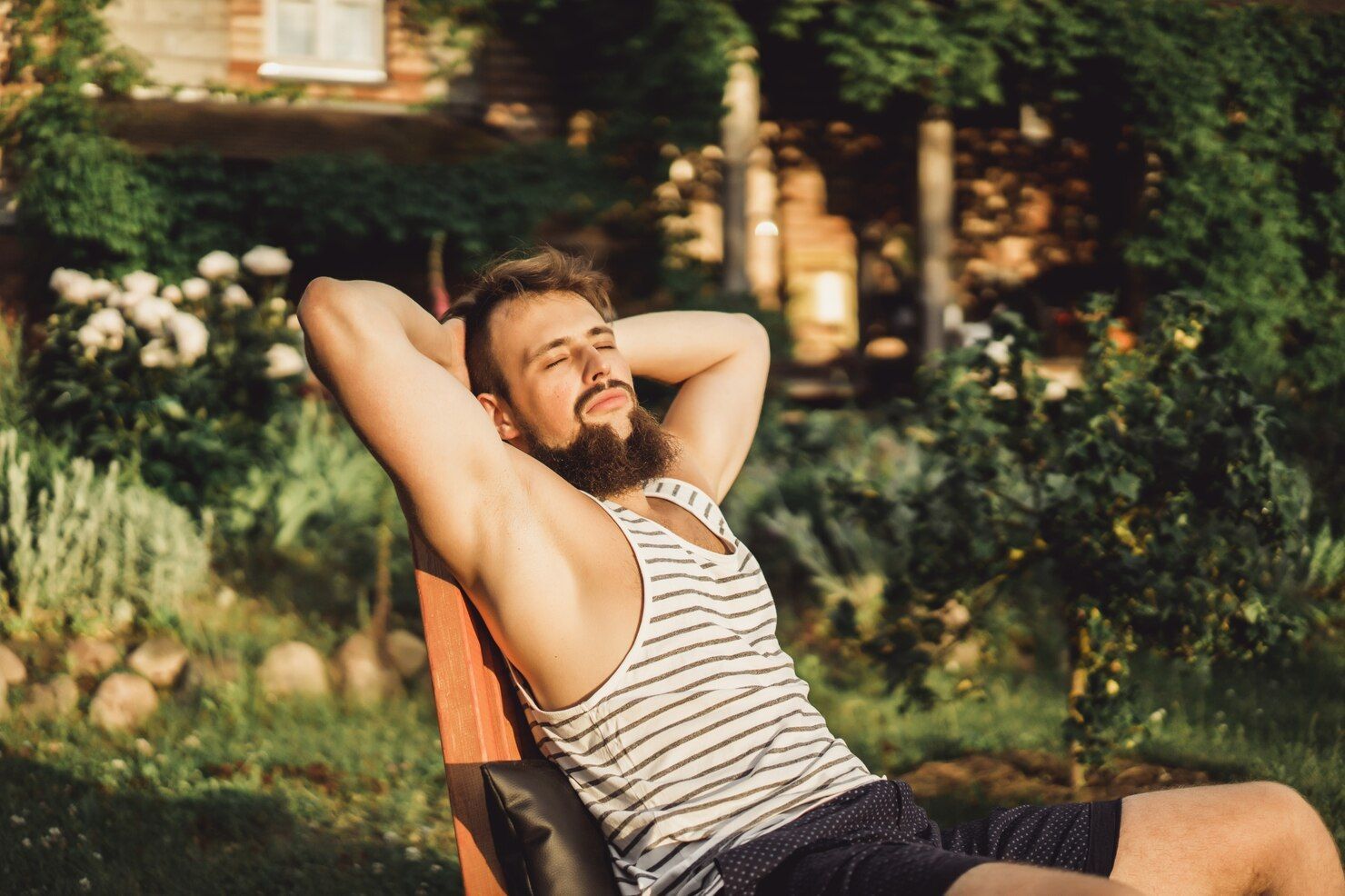 A man is sitting on a bench in a garden with his hands behind his head.
