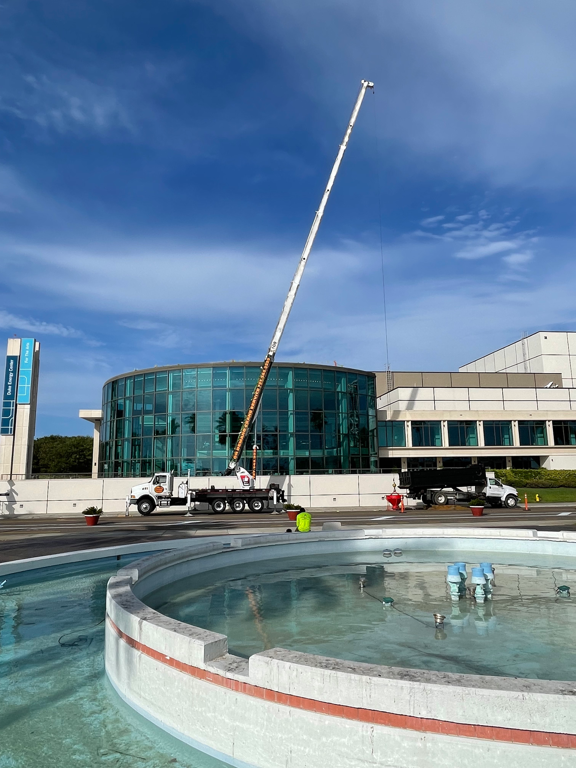 A tall crane near a round fountain and a modern building with glass windows under a blue sky.