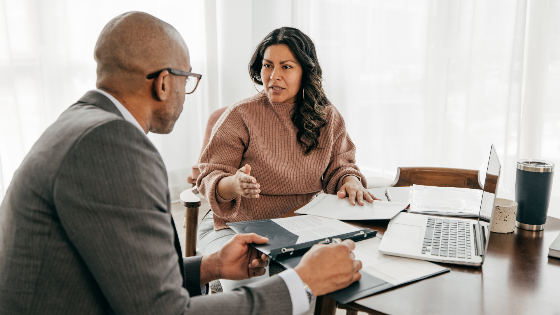 Man in glasses speaking, holding tablet, in meeting with blurred colleagues.