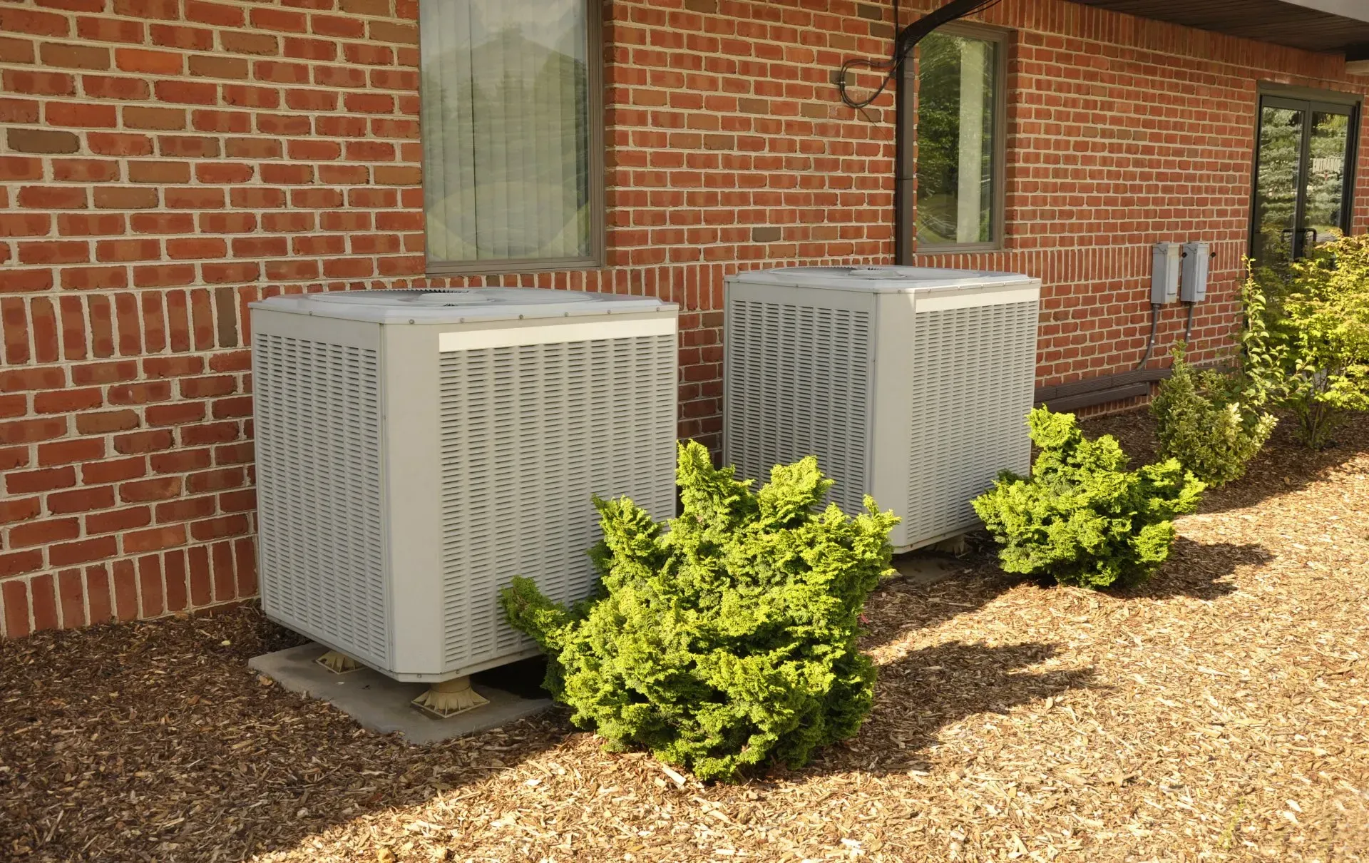 Two gray air conditioning units next to a brick building and green shrubs.