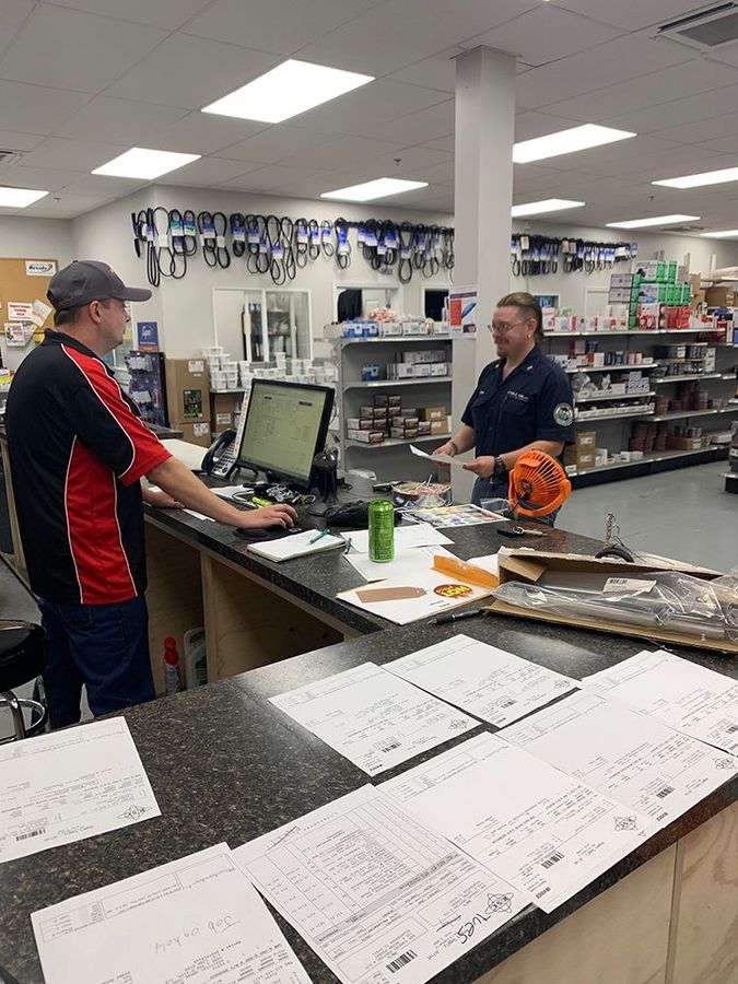 Two people at a store counter. One types on a computer, the other smiles, facing the other. Parts store interior.