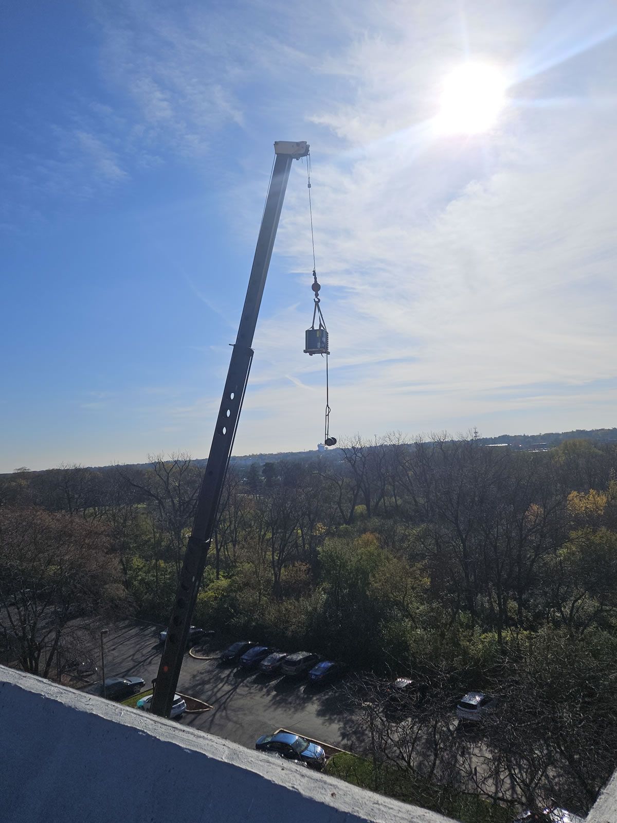 A crane lifting an object from a building rooftop on a sunny day. The object hangs mid-air over trees.