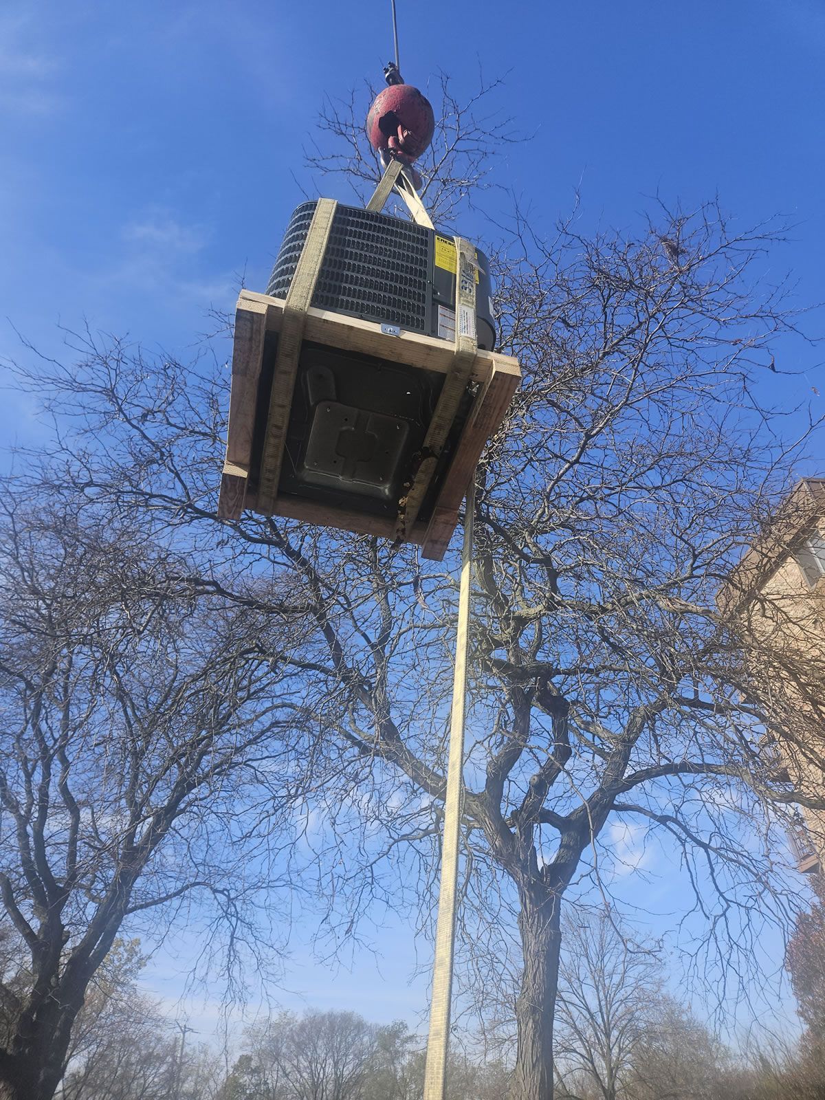 Crane lifting HVAC unit on wooden pallet near bare trees.
