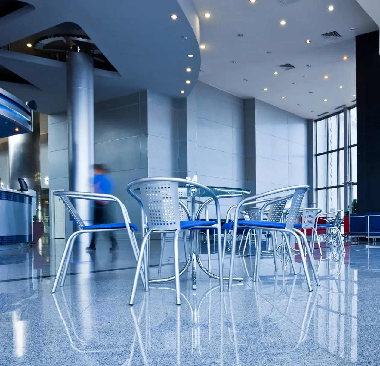 Modern lobby with chrome chairs, tables, reflective floor, and blurred figure walking past a counter.