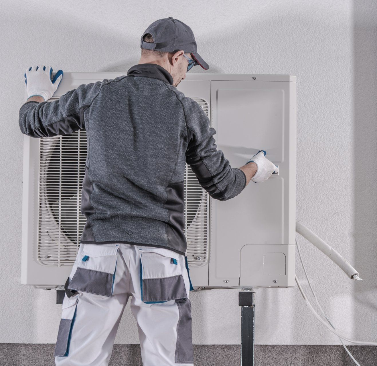 HVAC technician in gray and white work clothes installing an outdoor air conditioning unit on a white wall.