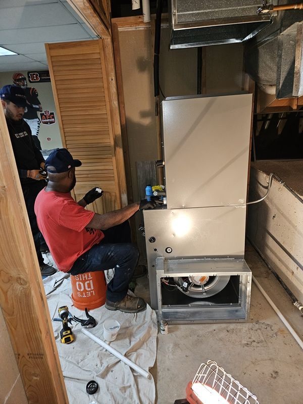 Two men work on a large HVAC unit in a basement, one in a red shirt and hat, the other in a blue shirt.