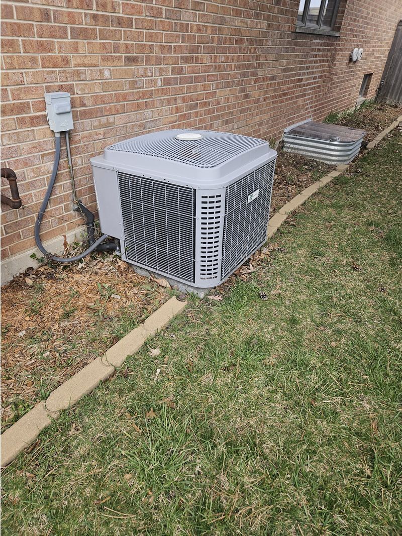Central air conditioning unit near a brick wall and a window well.