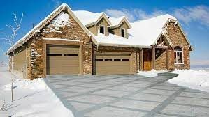 Stone house with snow-covered roof and driveway; two garage doors are closed; sunny winter day.