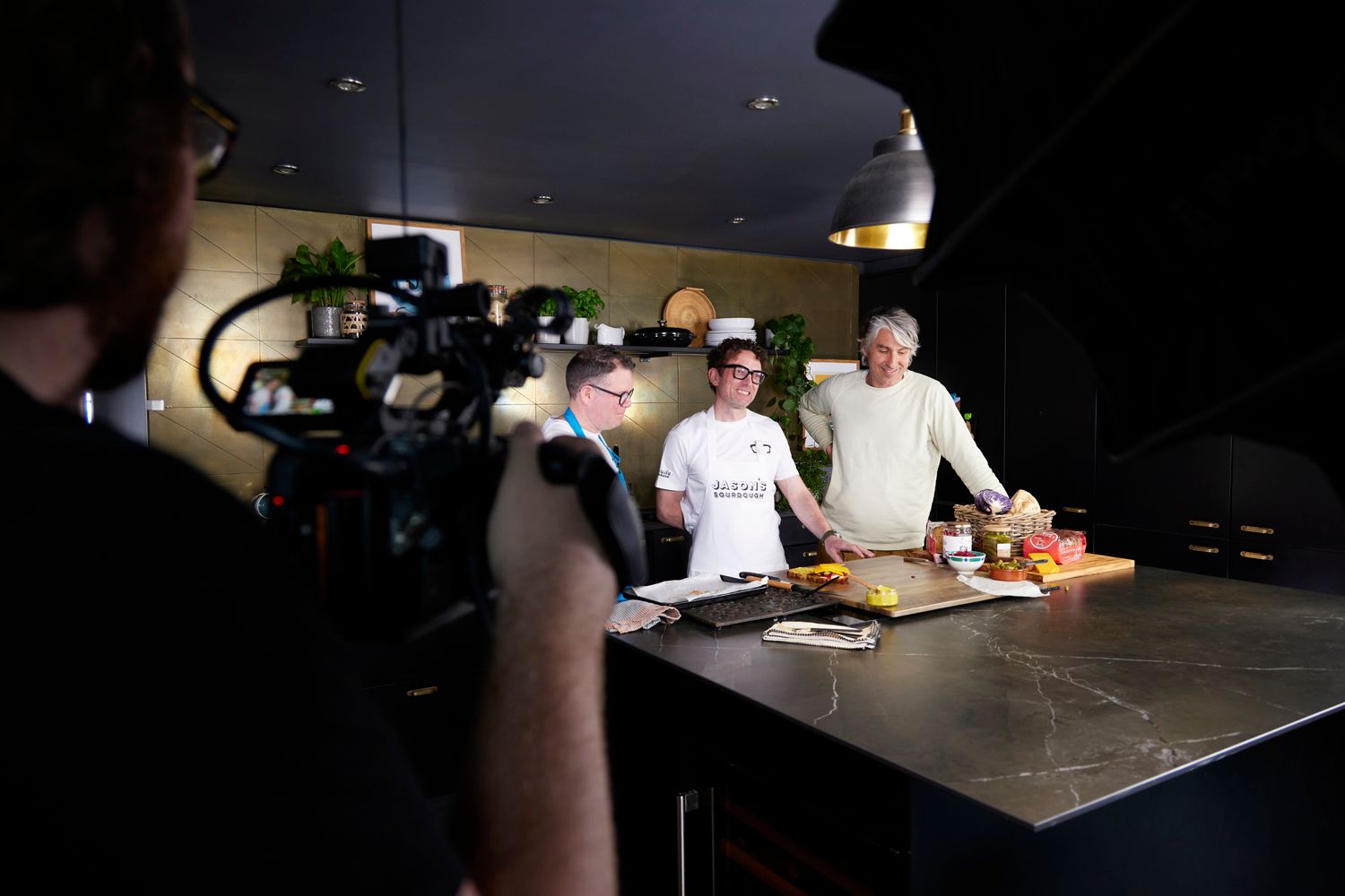 A man is holding a camera in front of a group of people in a kitchen.