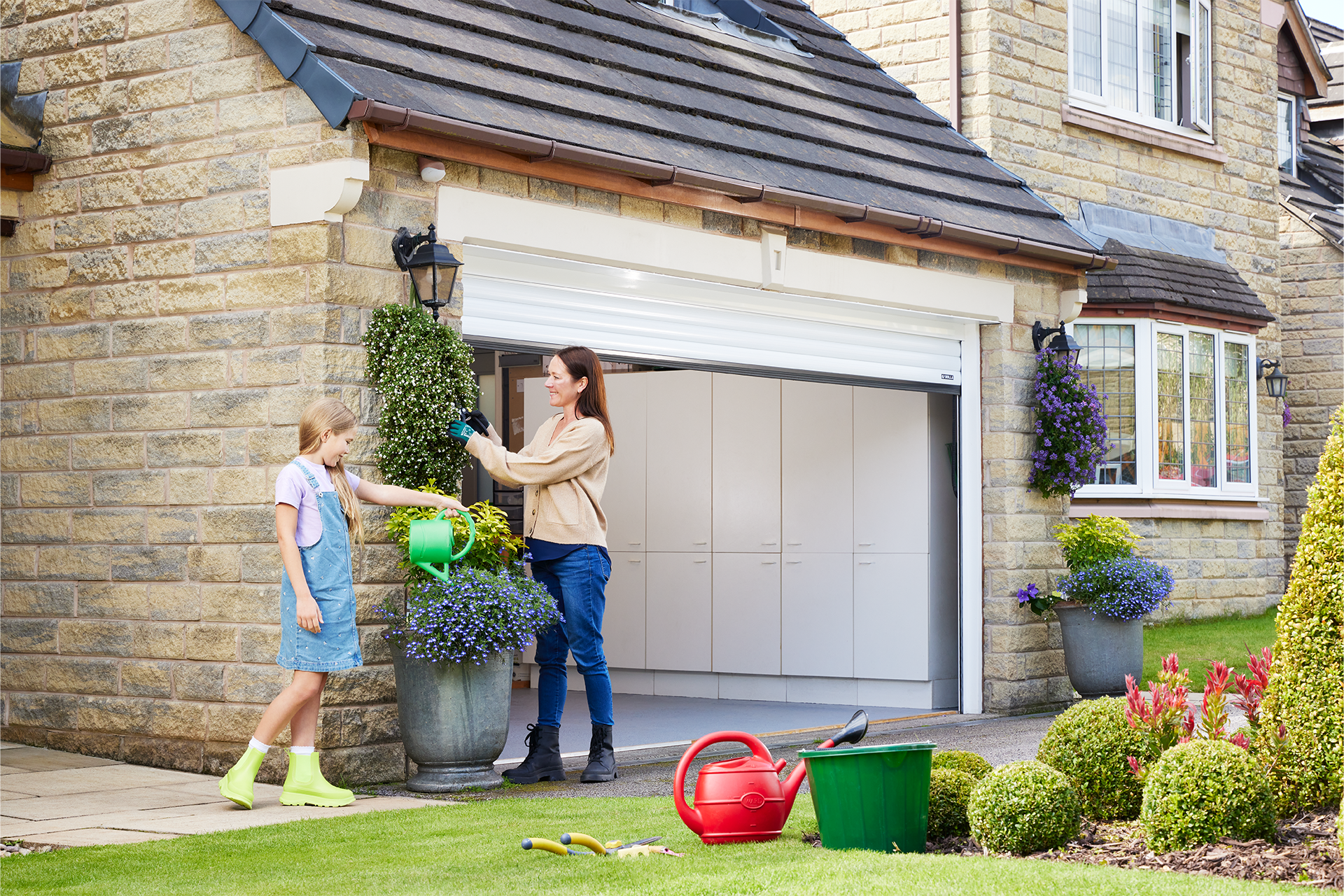 A woman and a girl are watering plants in front of a garage door.