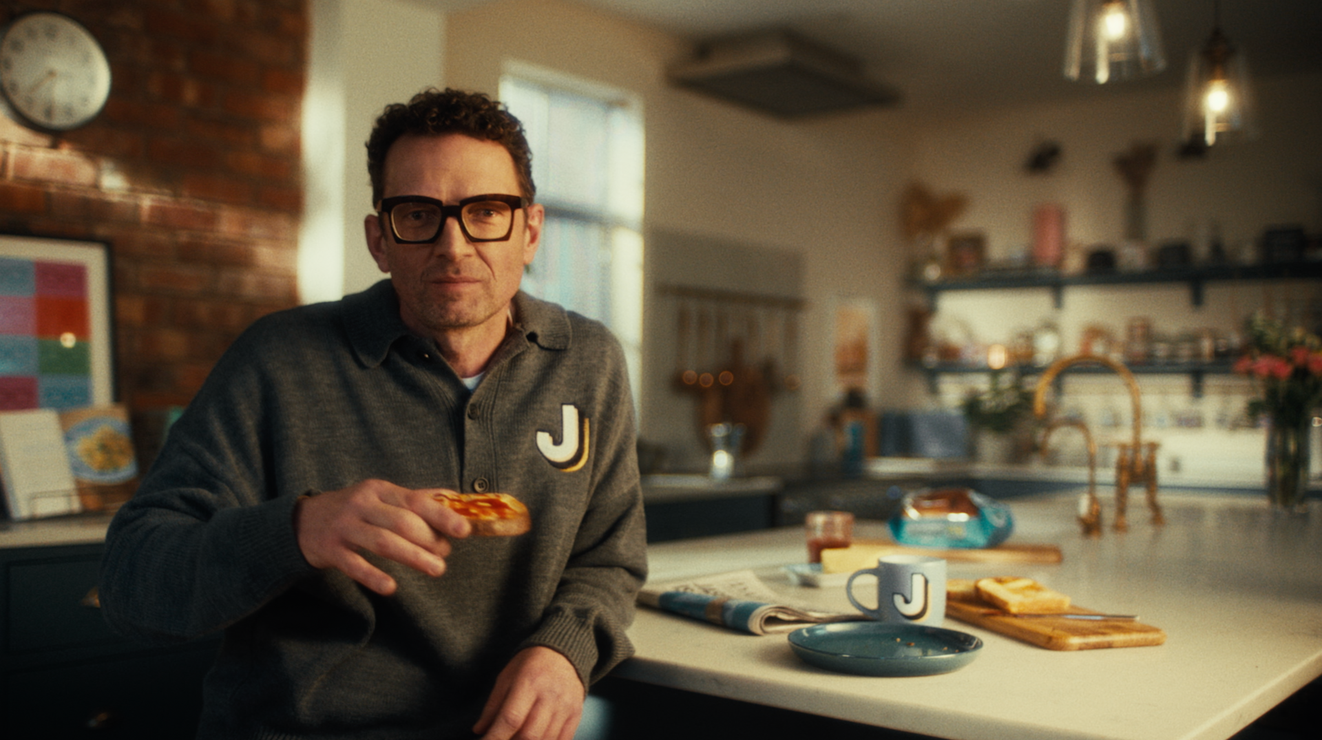 Man in glasses holding toast, sitting at a kitchen island. Kitchen setting, neutral tones, eating.