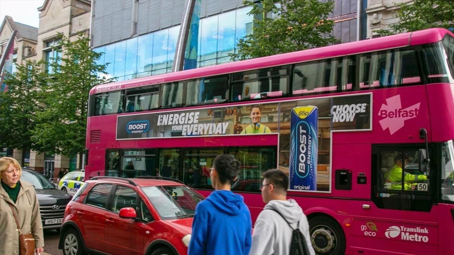 A pink double decker bus is parked in front of a building.