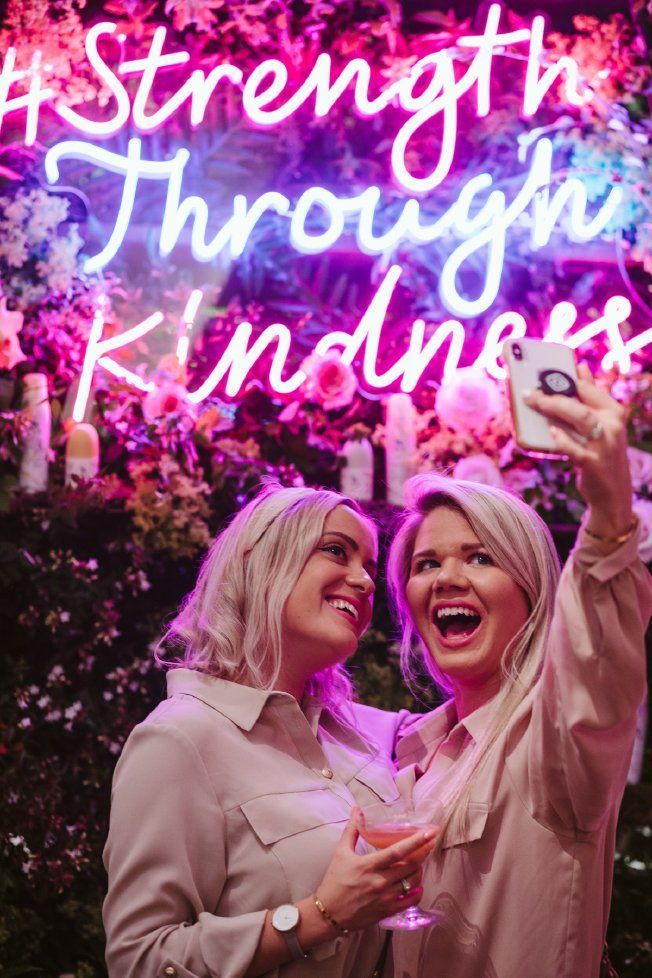 Two women are taking a selfie in front of a neon sign that says `` strength through kindness ''.