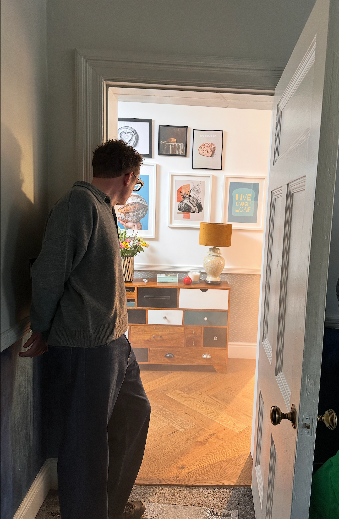 Man peers into a brightly lit room with framed artwork and a colorful cabinet through an open doorway.