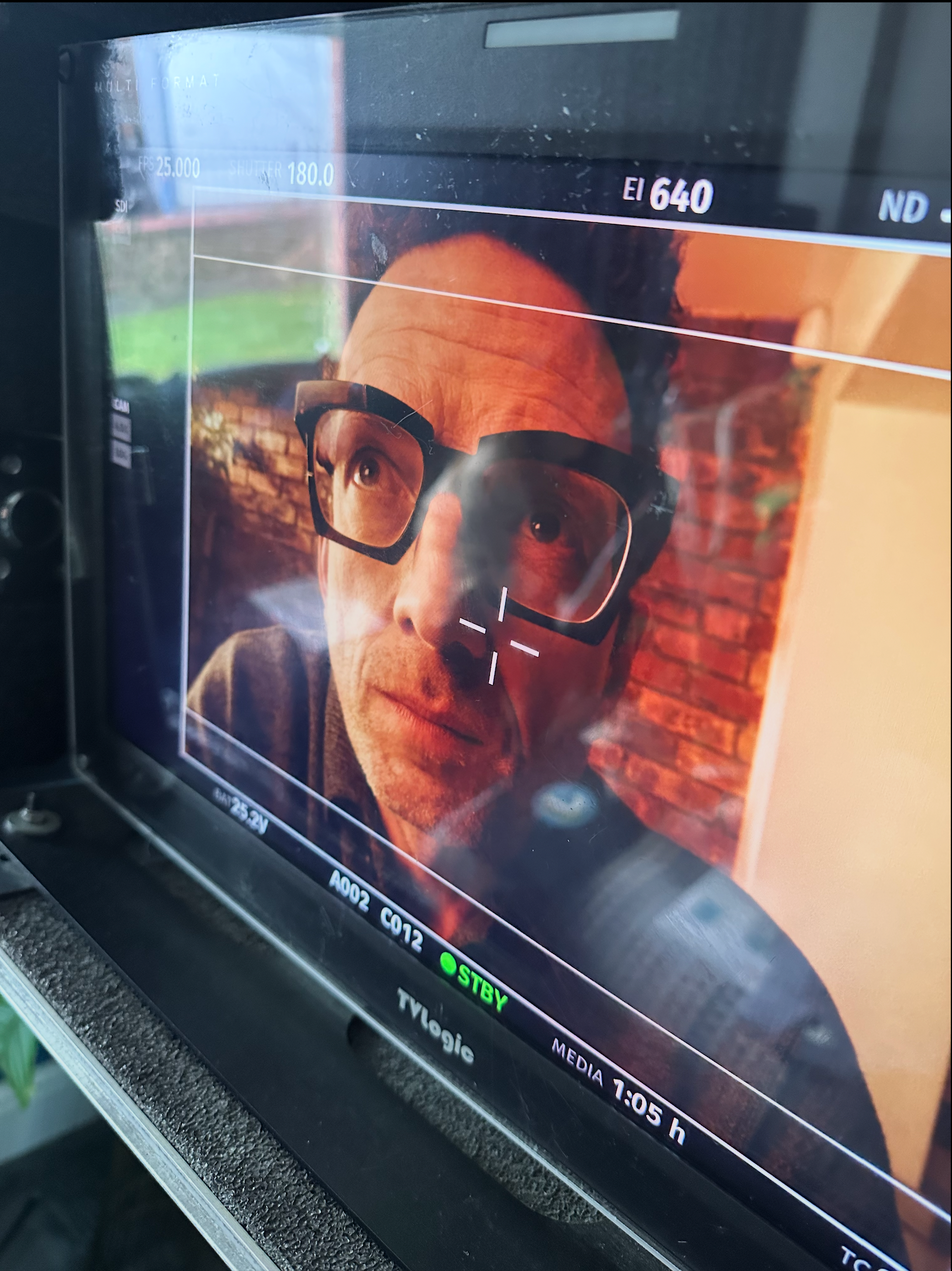 Man wearing glasses on a monitor screen, filming scene with brick wall in background.