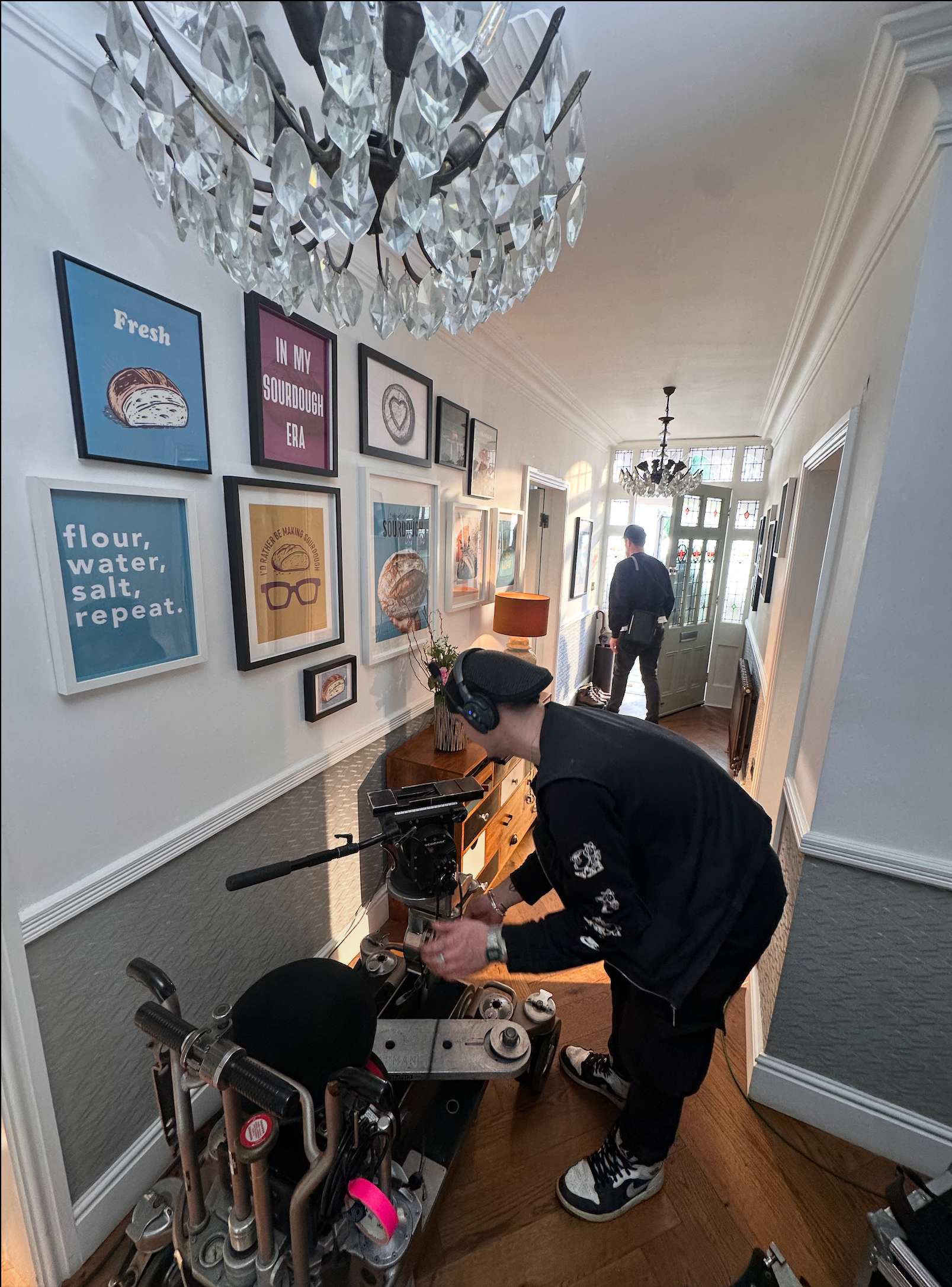 Person in black outfit adjusting a scooter in a hallway with framed art and chandelier.