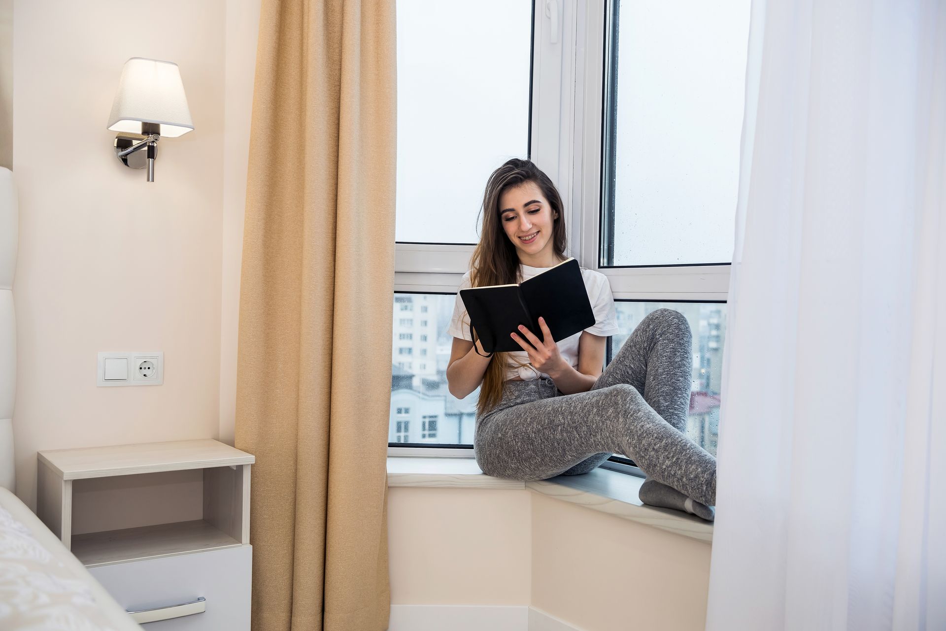 A woman is sitting on a window sill reading a book.