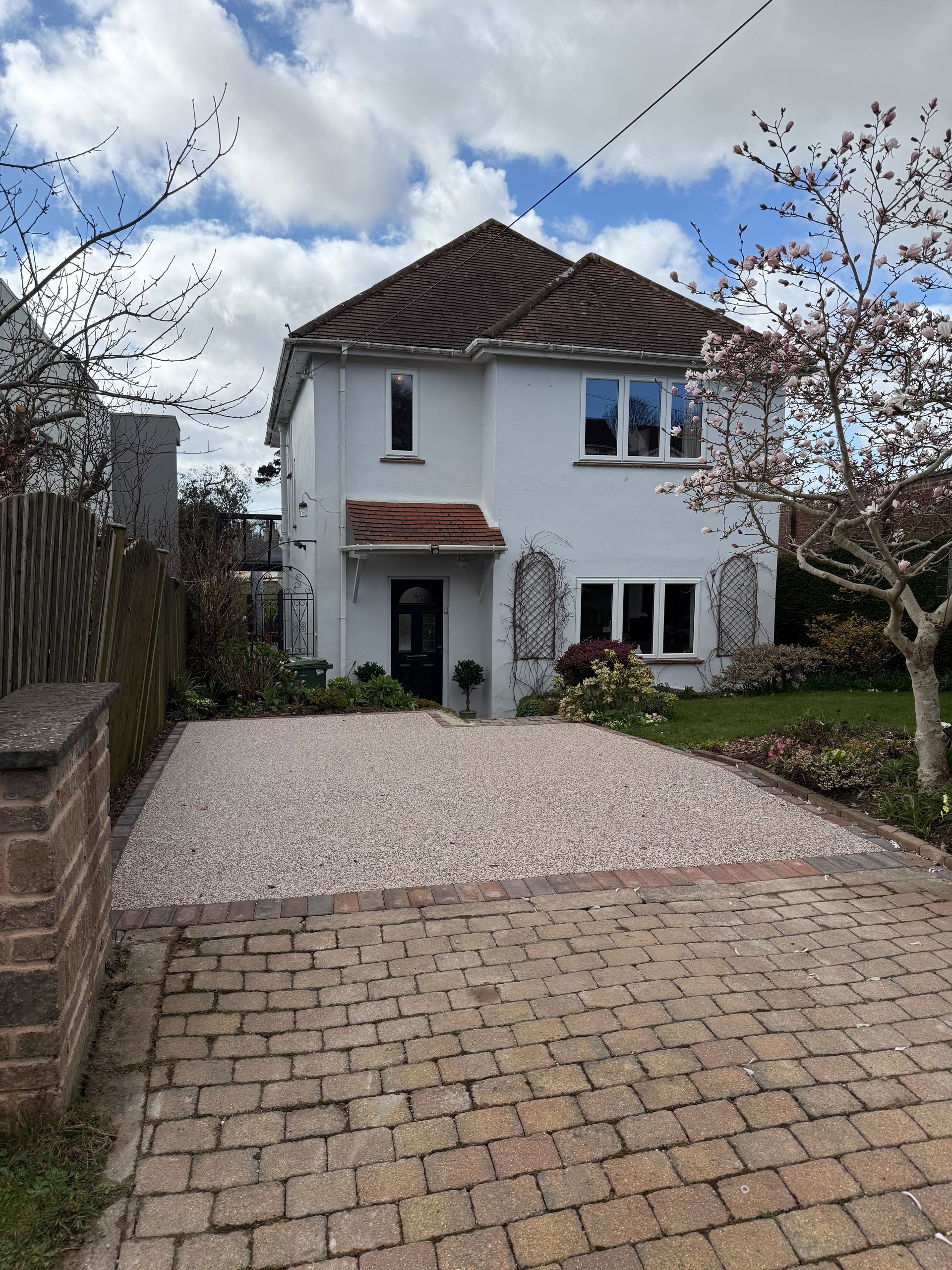 Two-story white house with brown roof, gravel driveway, brick pathway, and blooming tree.