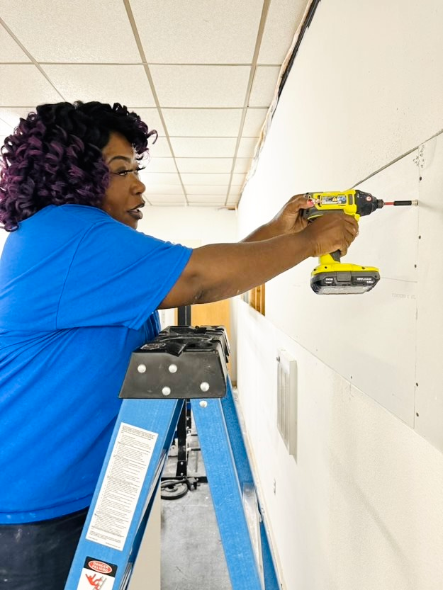 A woman is standing on a ladder using a drill on a wall.