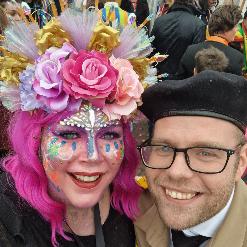 Twee lachende mensen in kleurrijke festivaloutfits en brillen poseren voor een selfie.