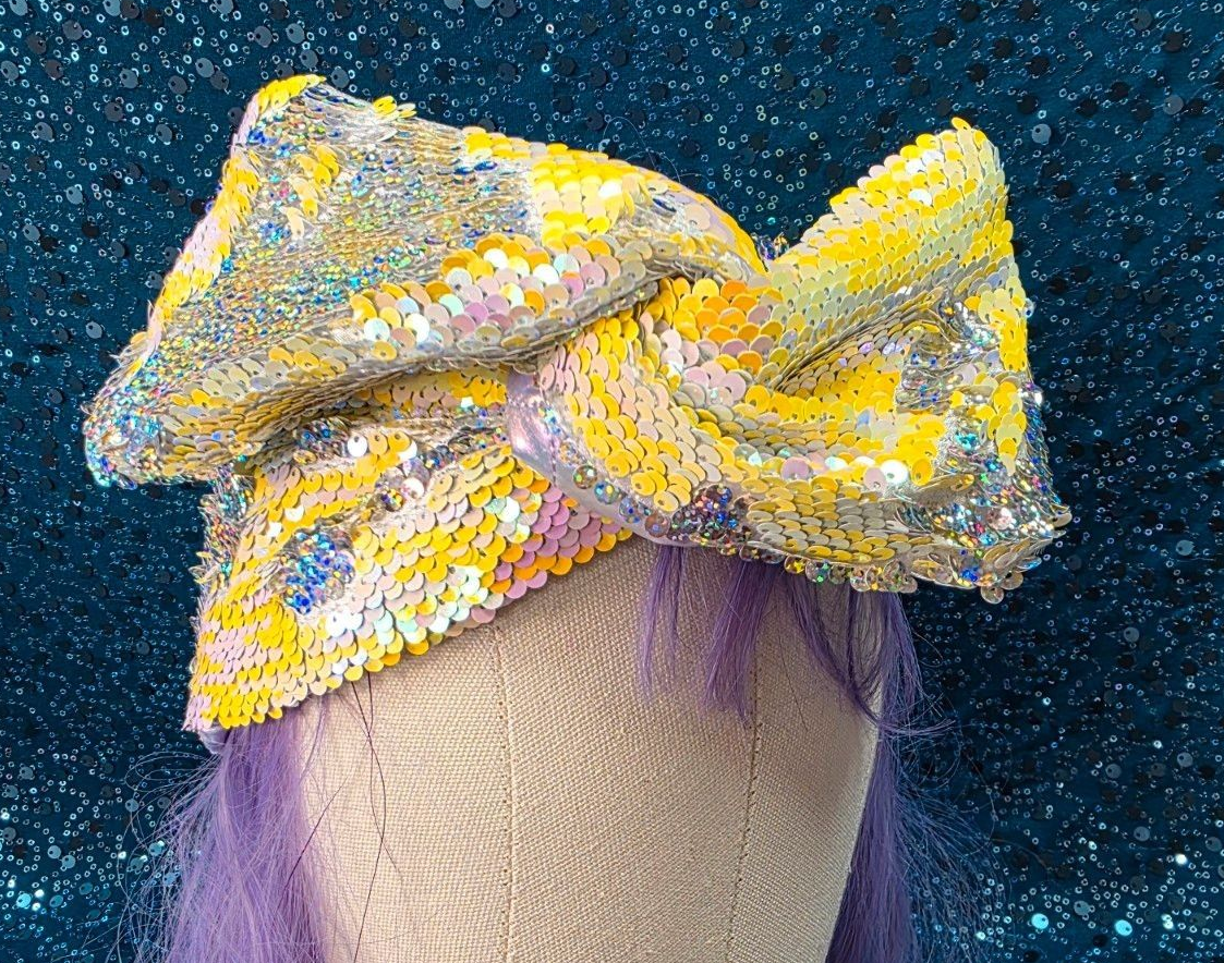Persoon met een grote geel-witte glitterwrap als hoofddeksel tegen een glinsterende blauwe achtergrond.