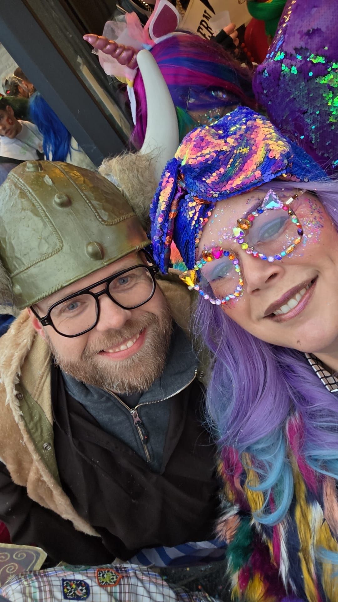 Twee lachende mensen in kleurrijke festivaloutfits en brillen poseren voor een selfie.