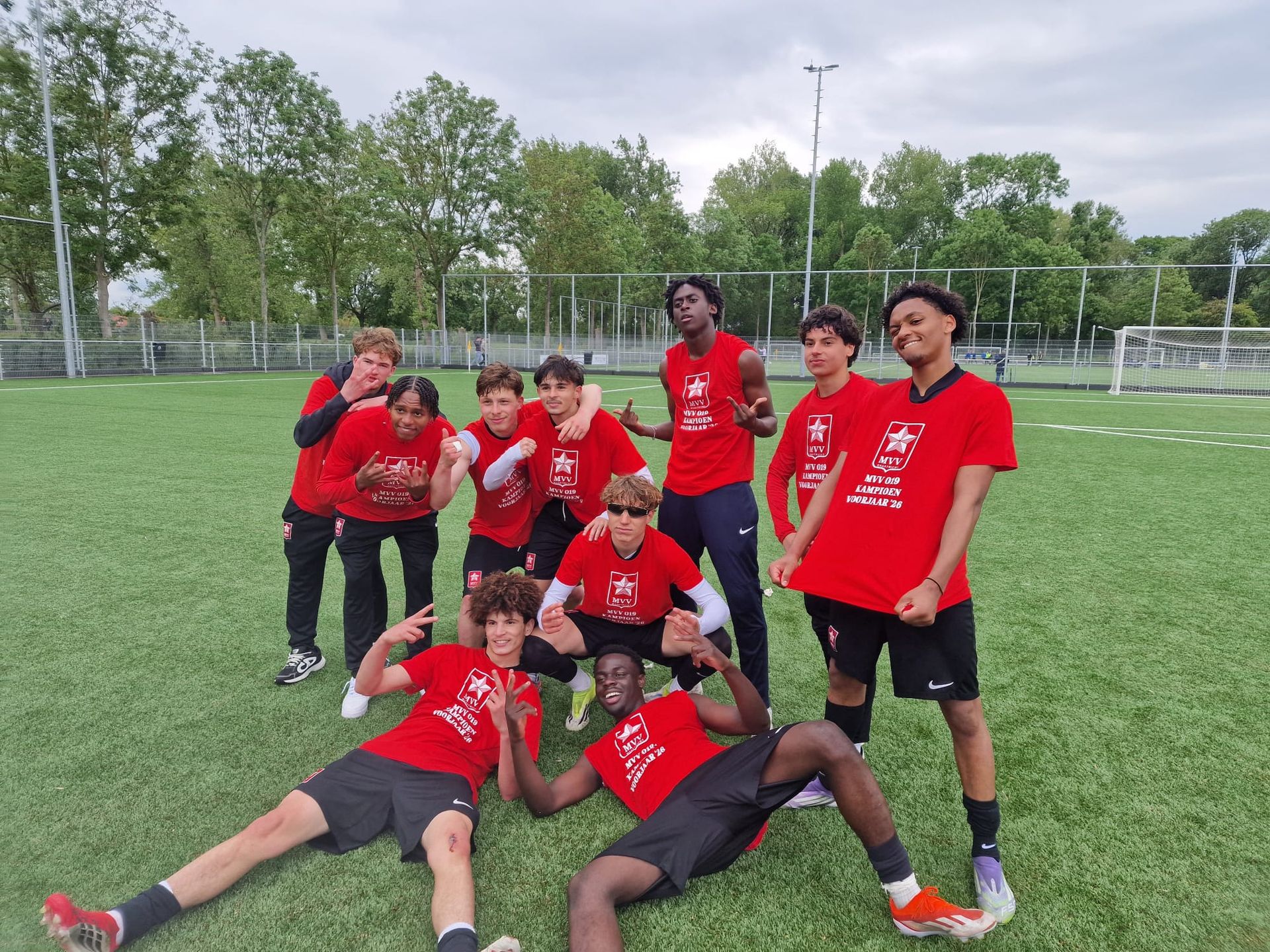 Een voetbalteam in rode shirts poseert op een veld, met bomen en doelpalen op de achtergrond.