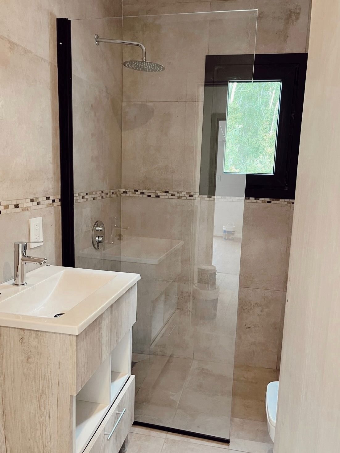 Baño moderno con ducha de cristal, lavabo con mueble, azulejos beige y ventana esmerilada.