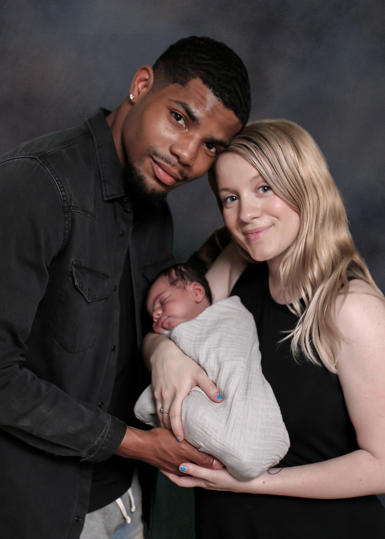 Couple gently holding a newborn wrapped in a white blanket against a dark studio backdrop