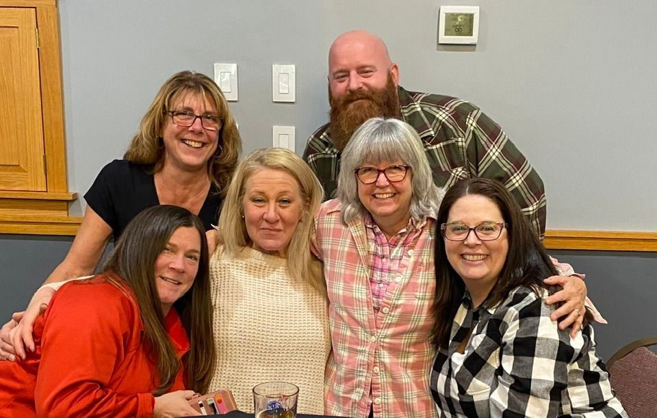 Six smiling people pose together indoors in front of a wooden wall.