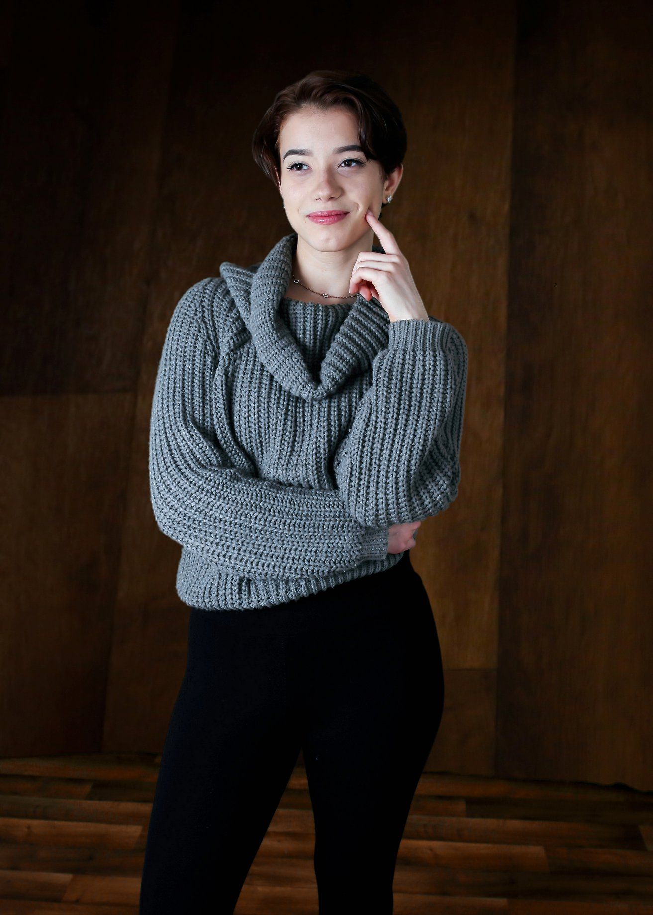 Person in a gray sweater and black pants stands against a dark wooden backdrop, hand near chin.