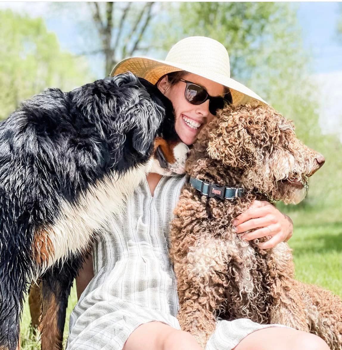 Woman in straw hat and sunglasses smiles as two dogs kiss her in a sunny outdoor setting.