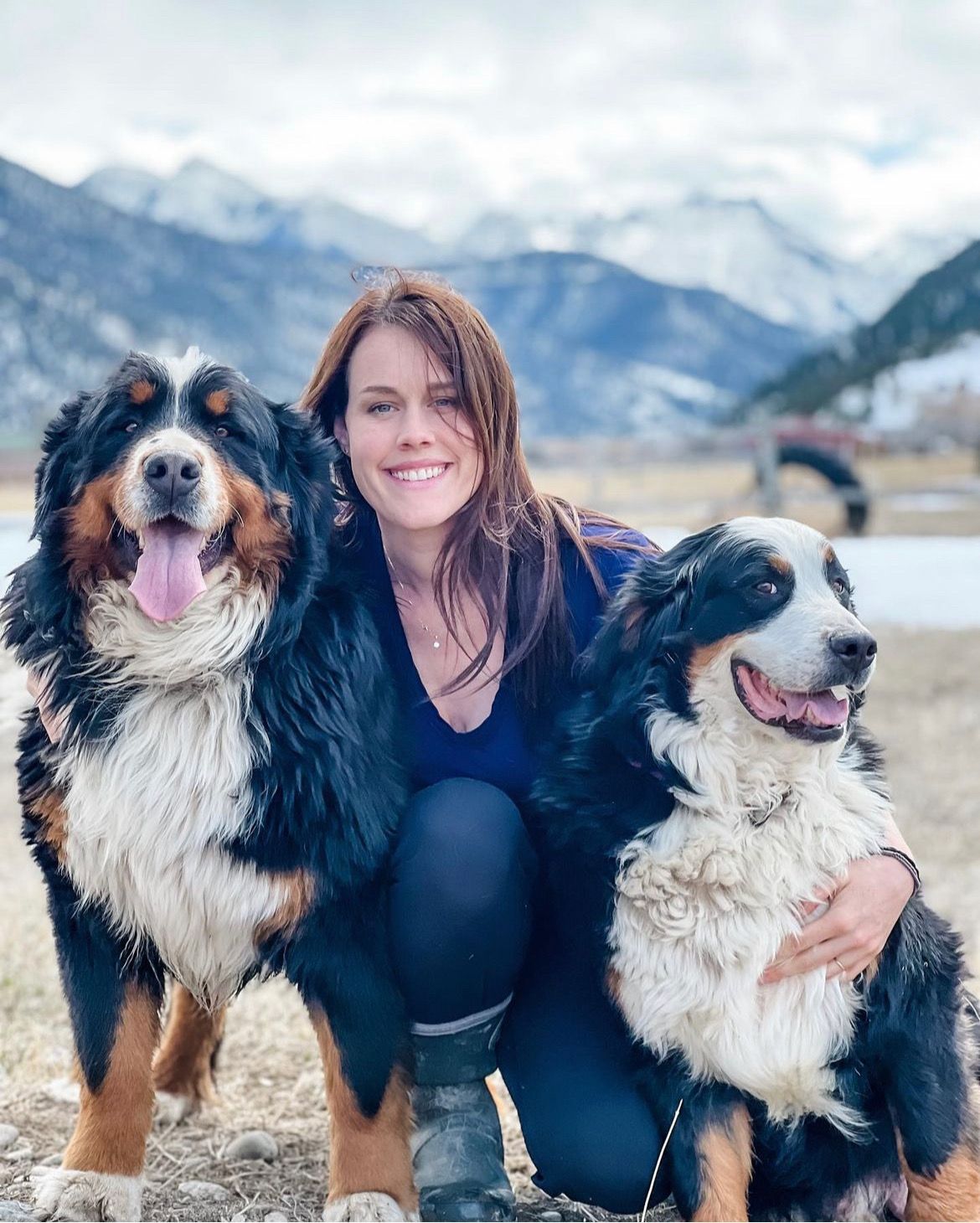 Woman kneels with two Bernese Mountain Dogs, smiles at camera; snowy mountains in background.