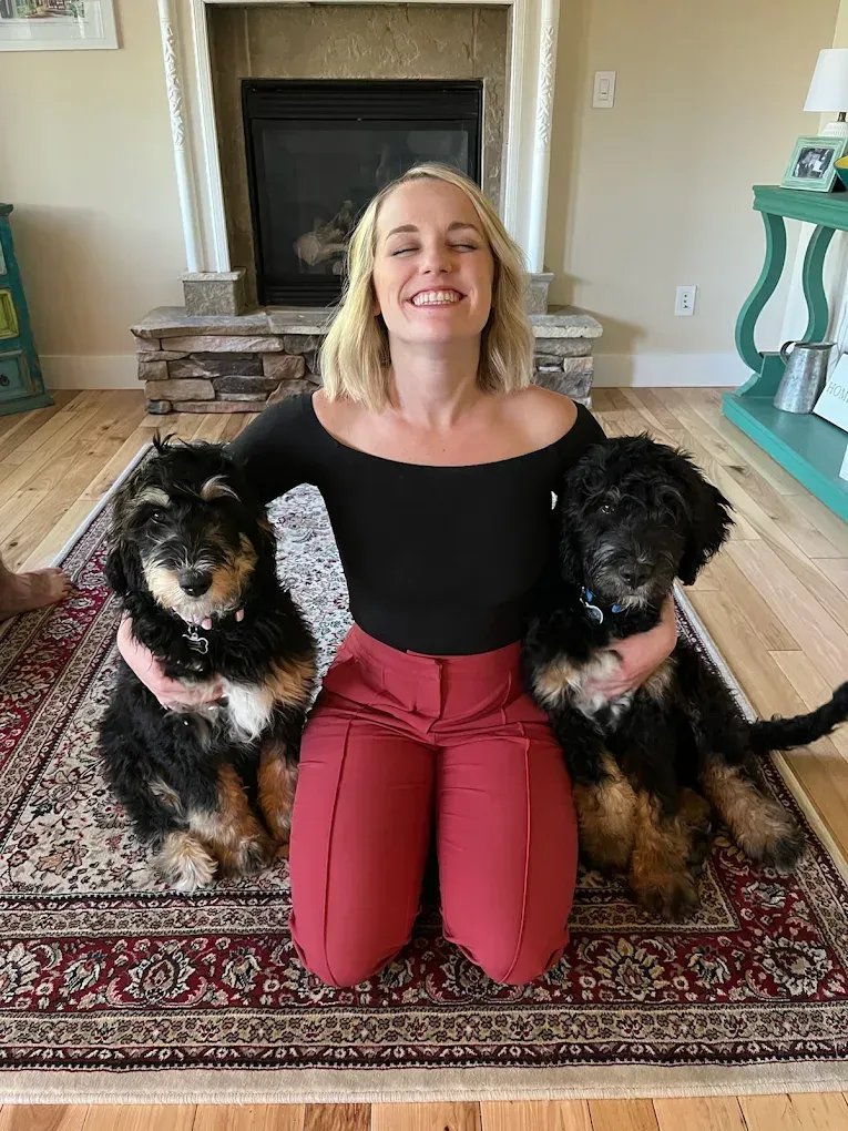 Woman kneels with two Bernedoodle puppies. She smiles broadly, wearing a black top and red pants on a rug near a fireplace.