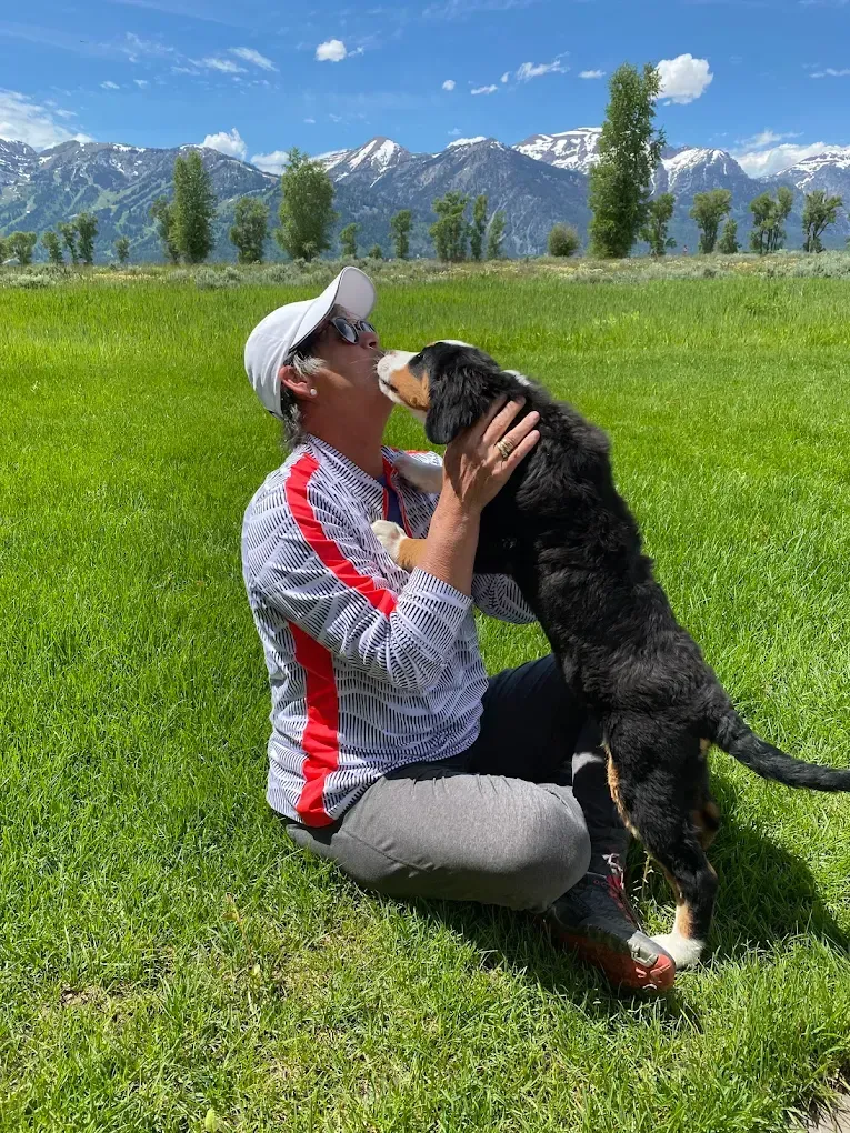 Woman in white hat and grey sweats hugging a black and brown dog on a green lawn, mountains in background.