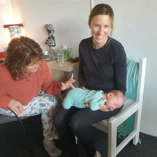 Two women sit with a sleeping baby in a chair; one holds the baby while the other looks on.