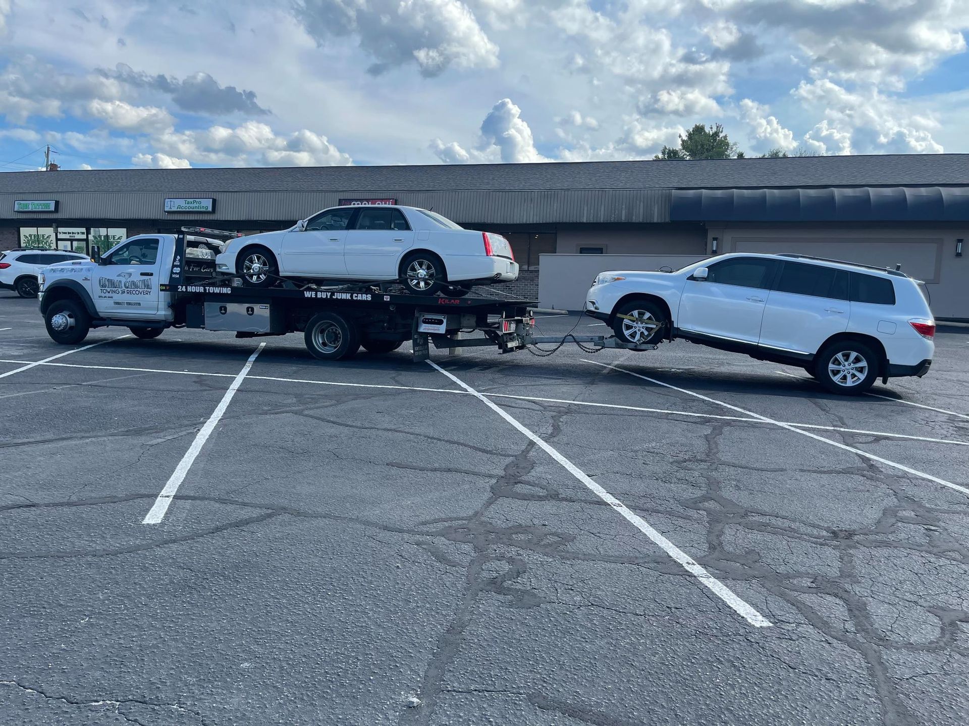 A tow truck is towing two cars in a parking lot.