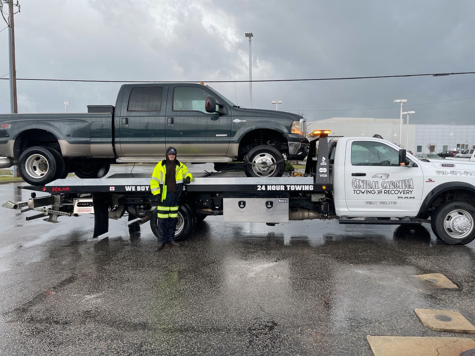 Tow truck carrying a dark green pickup truck; person in safety vest stands near it on wet pavement.