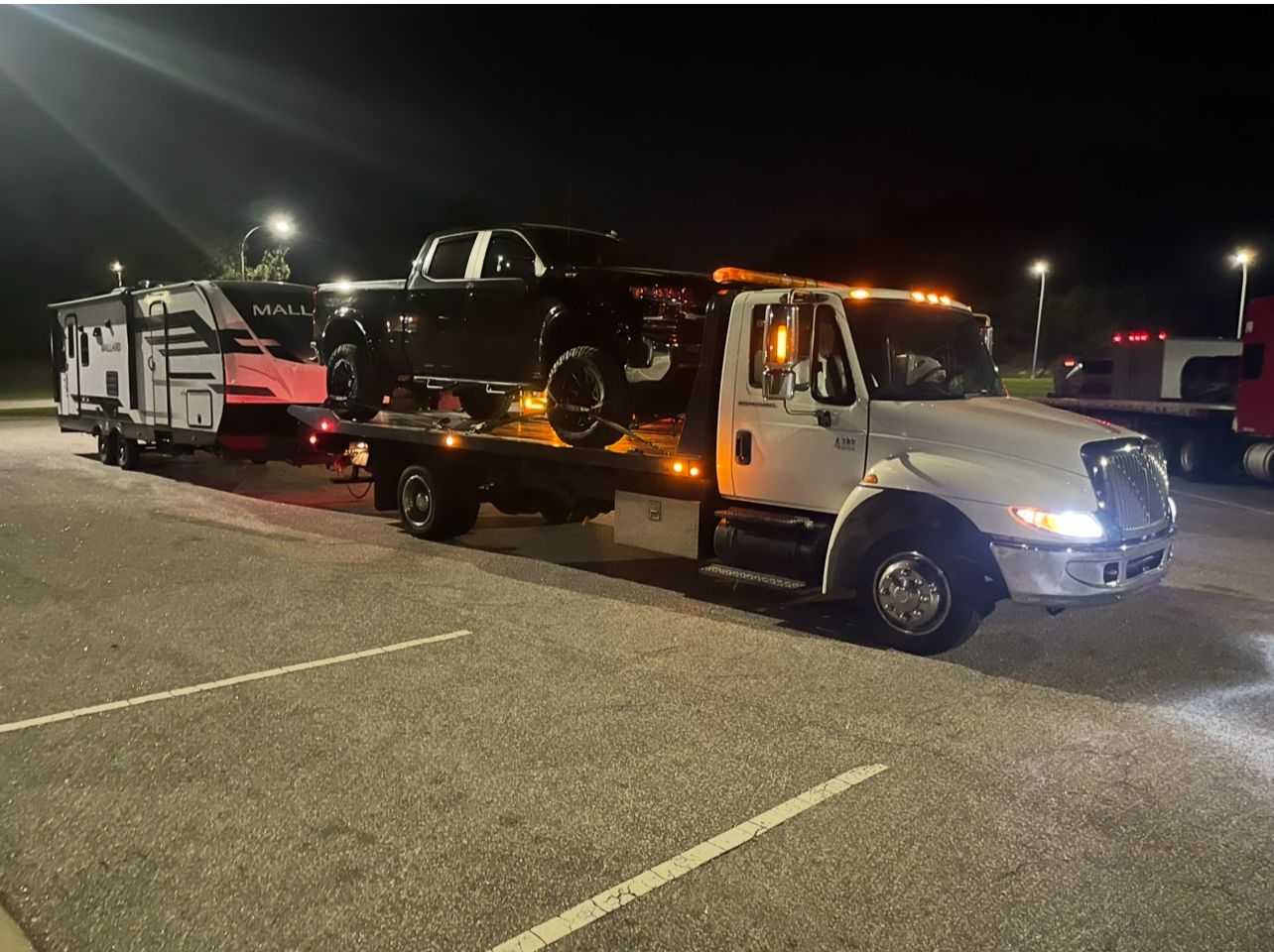A tow truck is towing a truck and trailer in a parking lot at night.