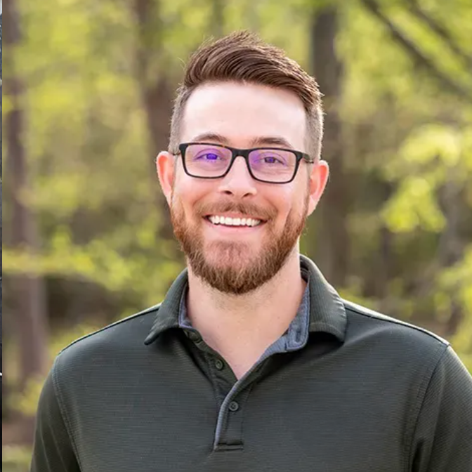 Smiling man with glasses and beard, wearing a green polo shirt, outdoors.