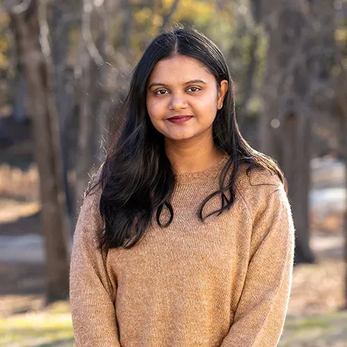 A woman in a brown sweater is standing in front of a forest.