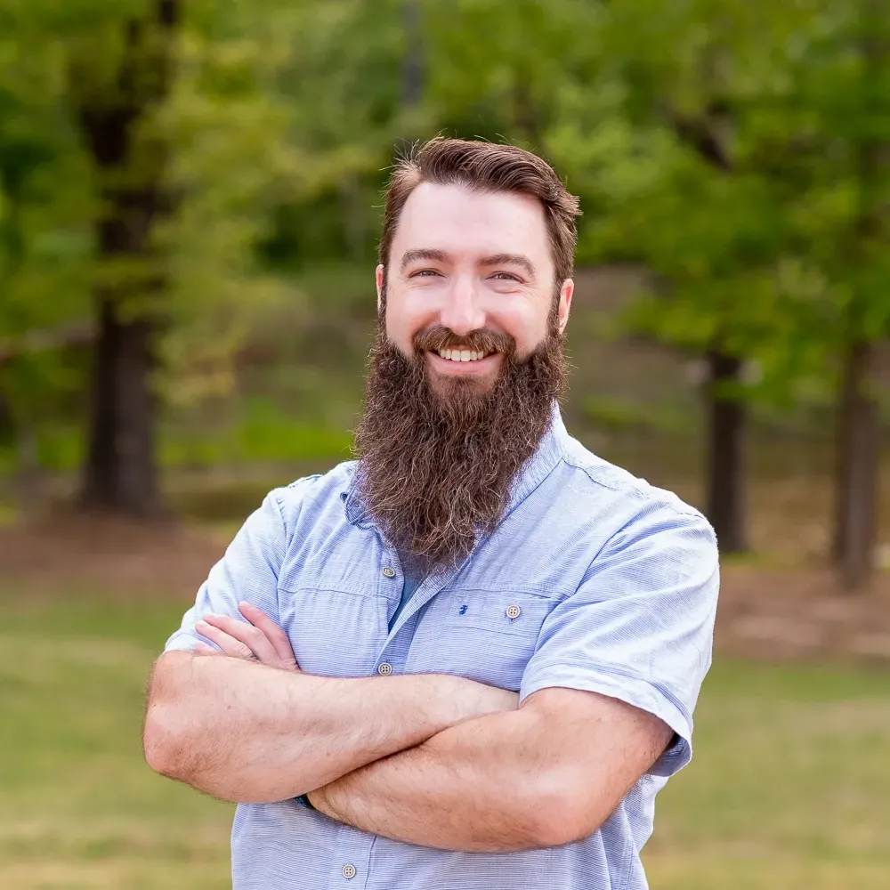 A person with a long beard and light blue collared shirt smiling with arms crossed, set against a blurry park background.