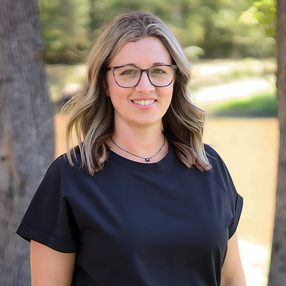A woman wearing glasses and a black shirt is standing in front of a tree.