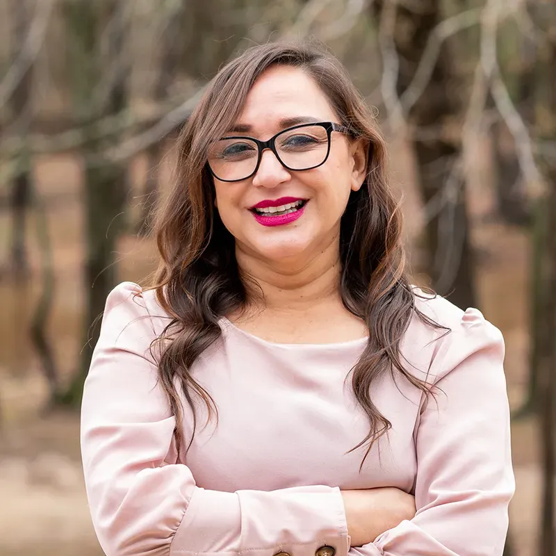 A woman wearing glasses and a pink shirt is smiling with her arms crossed.