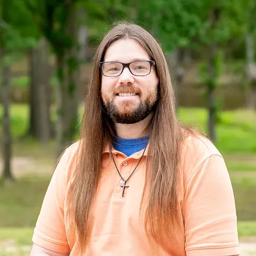 A man with long hair and a beard is wearing glasses and a cross necklace.