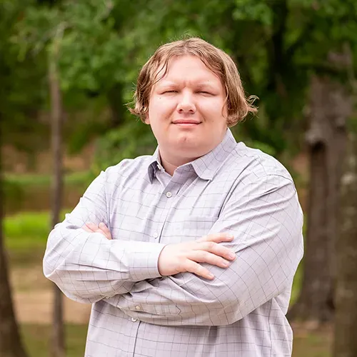 Man with arms crossed, wearing a button-down shirt, standing outdoors, trees in background.